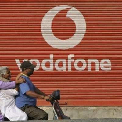 A man and two passengers ride on a scooter past a shop displaying the Vodafone logo on its shutter in Jammu