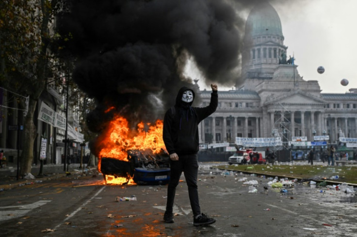 A demonstrator wearing a Guy Fawkes mask walks in front of a Cadena 3 radio station car set on fire during a protest outside the National Congress in Buenos Aires